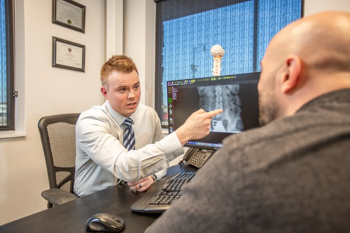 Chiropractor pointing to a spinal X-ray on a monitor while consulting with a patient