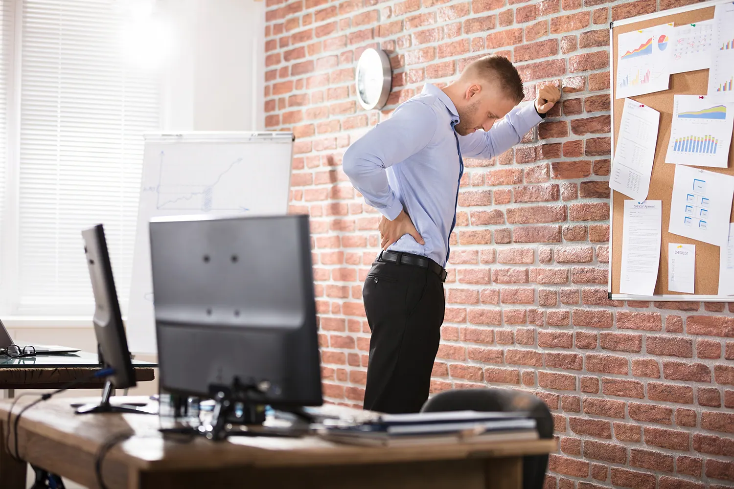 Man in an office holding his lower back while leaning on a brick wall