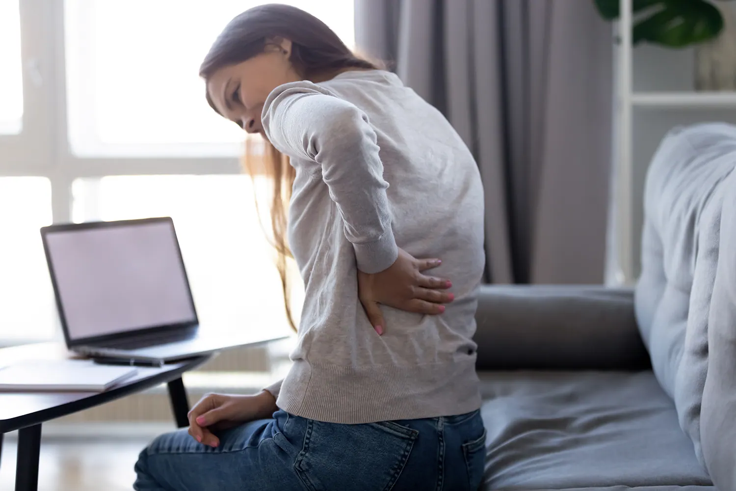 Woman sitting on a sofa with a laptop, holding her lower back